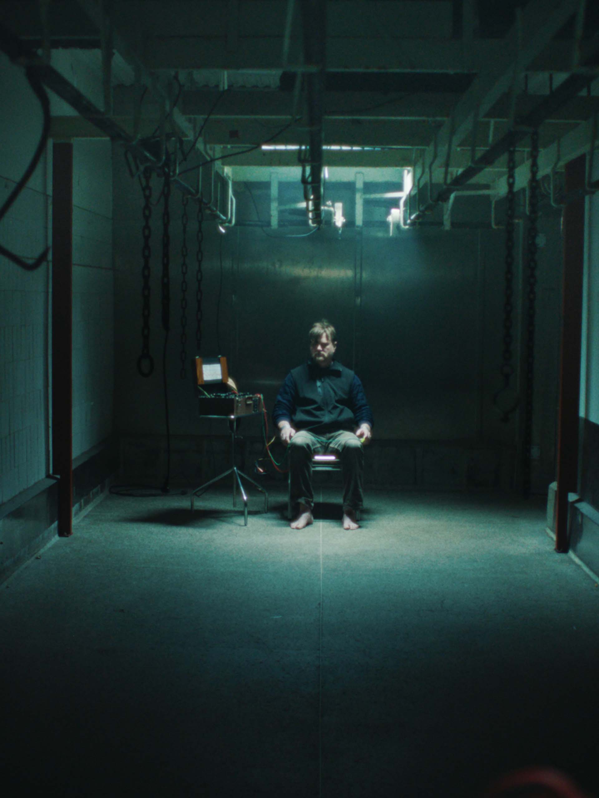 A barefoot man sitting in a dimly lit, industrial room with chains hanging from the ceiling and connected to a device on a stand beside him.