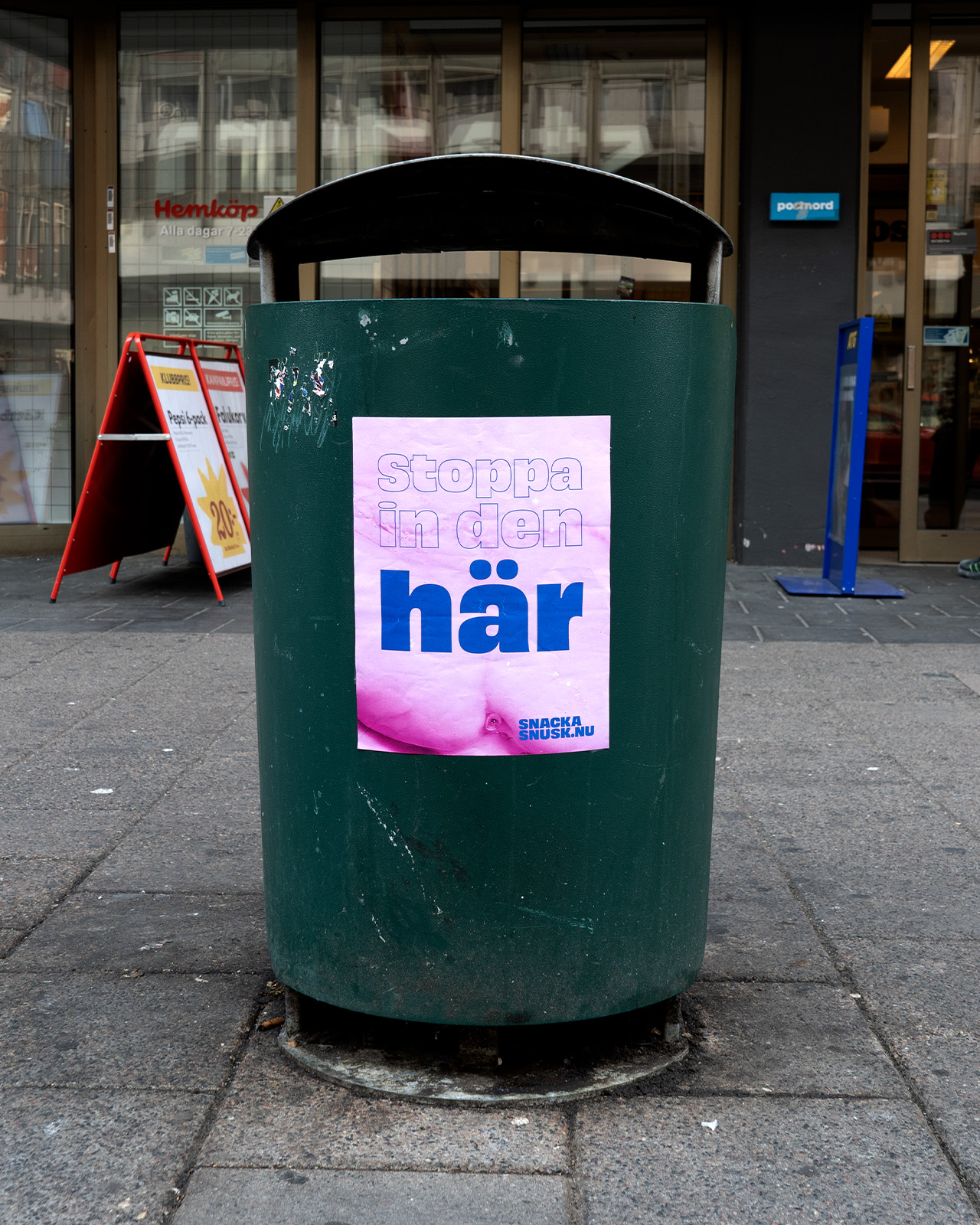 Green trash bin on a city sidewalk with a pink poster displaying Swedish text 'Stoppa in den här'.