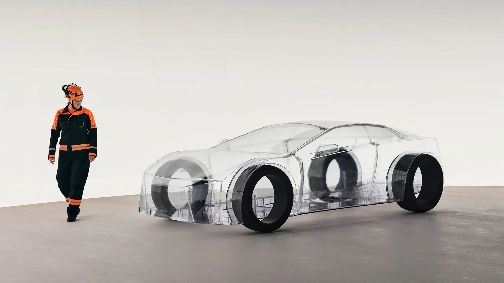 Person in orange and black protective gear walking beside a transparent car model with visible internal structure and large tires.