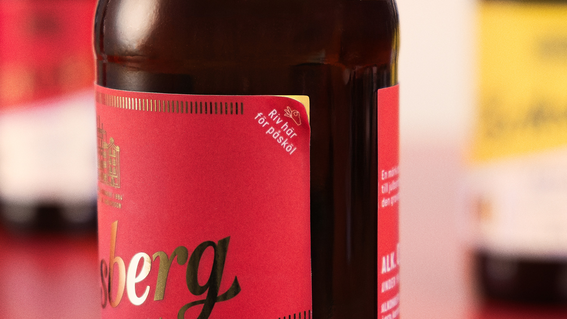 Close-up of a brown glass bottle with a red label showing gold text and a tear strip marked 'Riv här för påsköl'.
