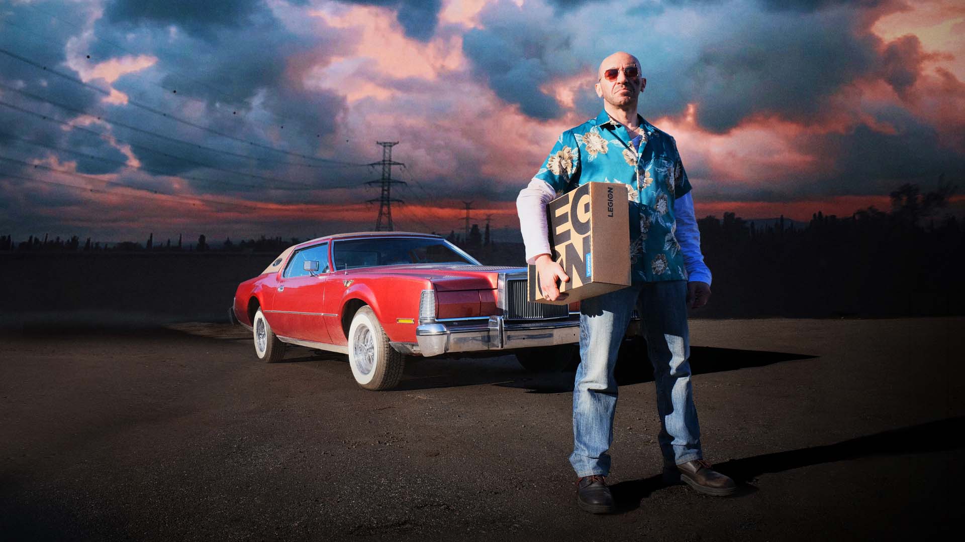 Man wearing sunglasses, a blue floral shirt, and jeans stands holding a Lenovo Legion box in front of a red vintage car at sunset.