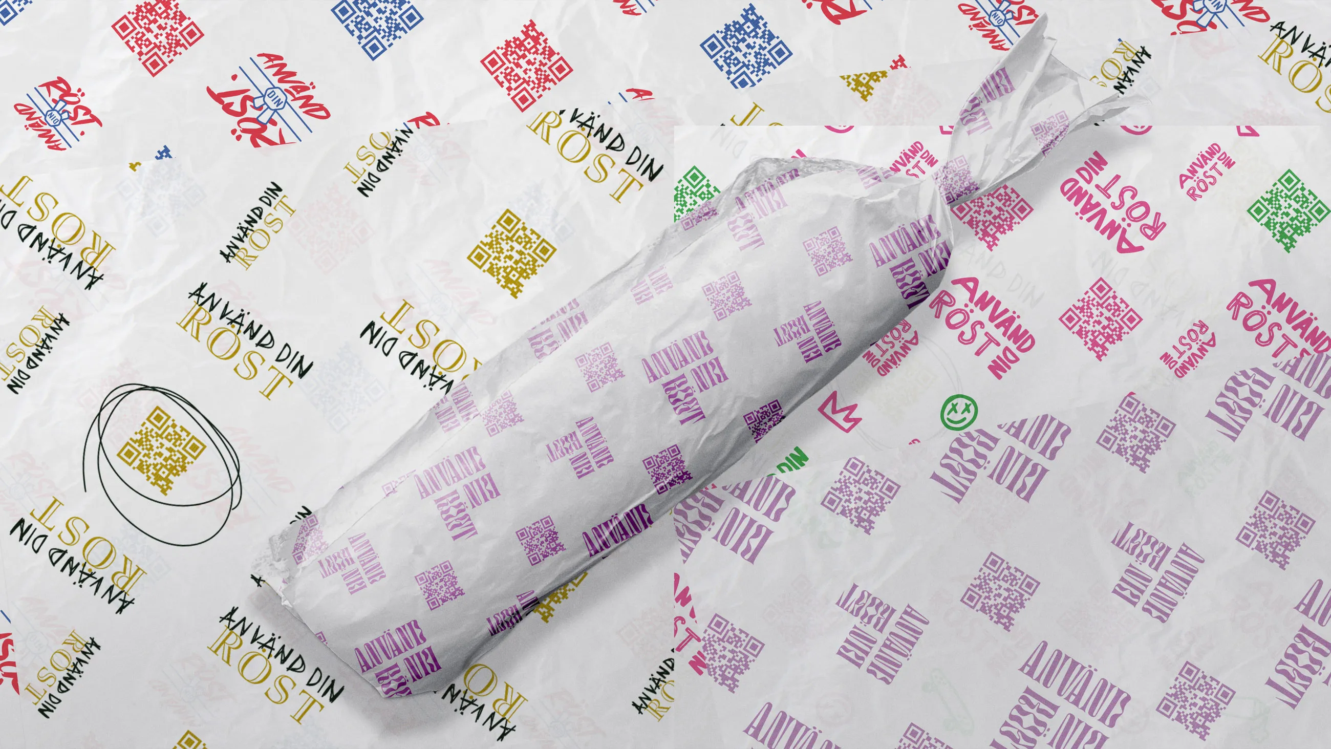 Wrapped sandwich in white paper with purple QR codes and 'ANVÄND RÖST' text, resting on a surface with multicolored QR codes and 'ANVÄND DIN RÖST' repeated.