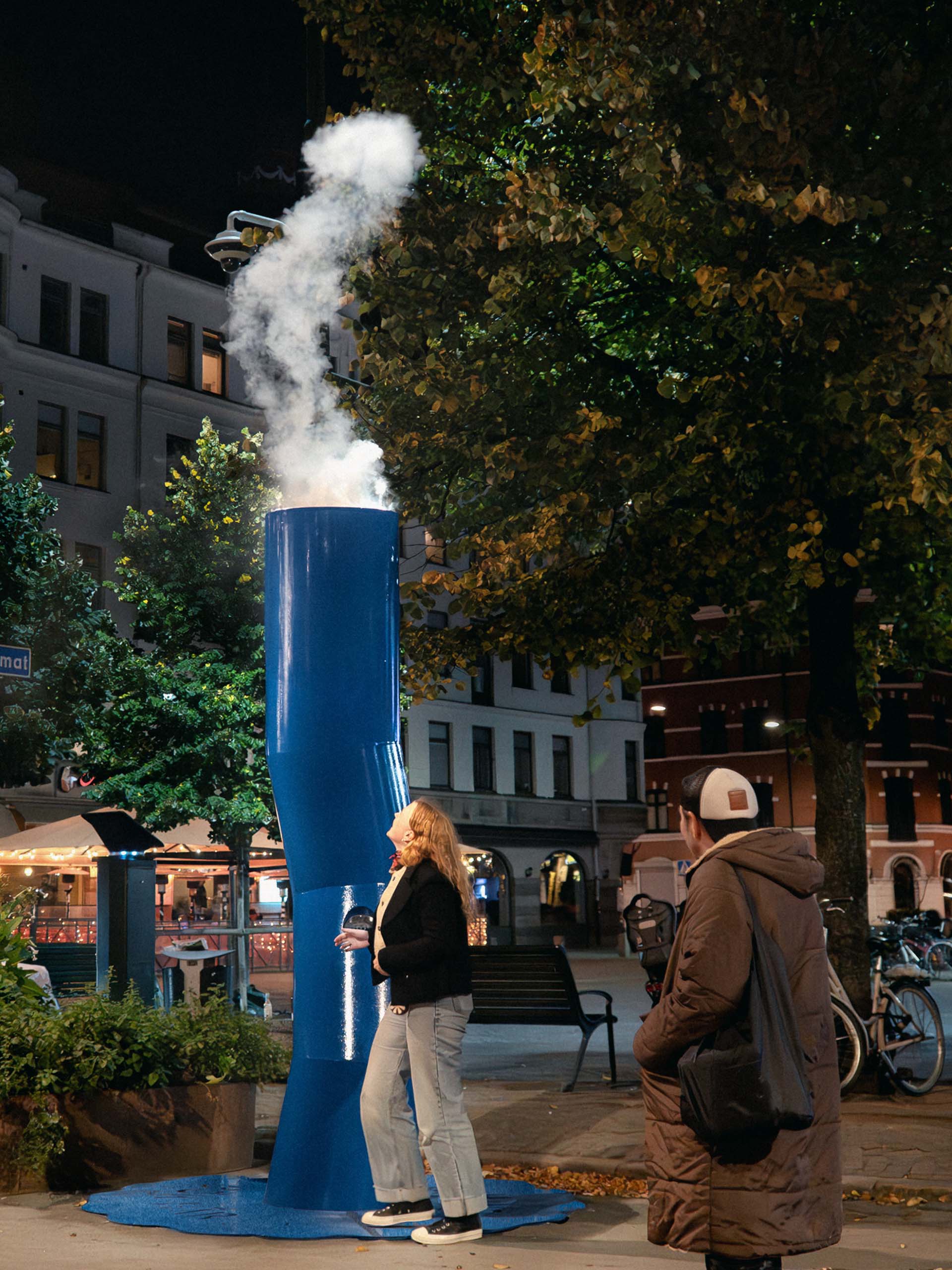 Woman standing near a tall blue sculpture emitting smoke at night, with a man watching nearby in an urban park.