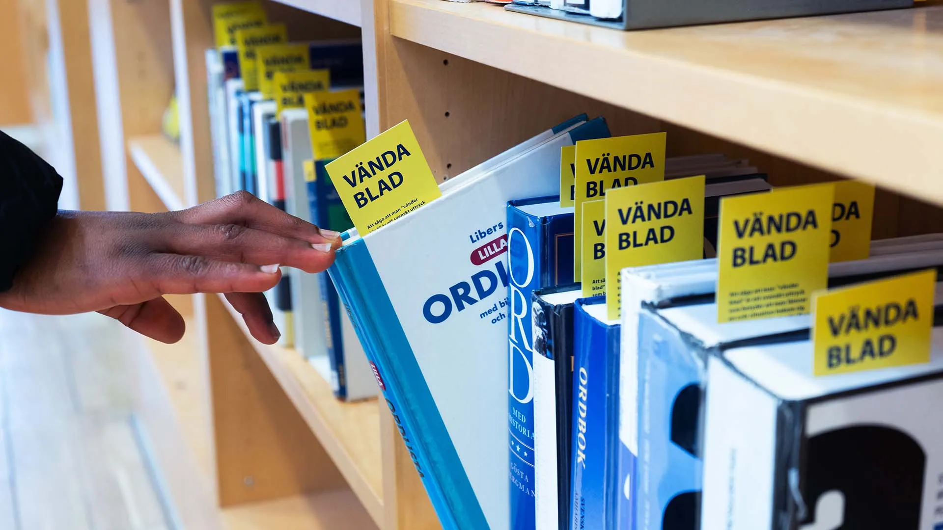 Hand pulling a blue and white book from a shelf with yellow bookmarks labeled 'VÄNDA BLAD' in a library or bookstore.