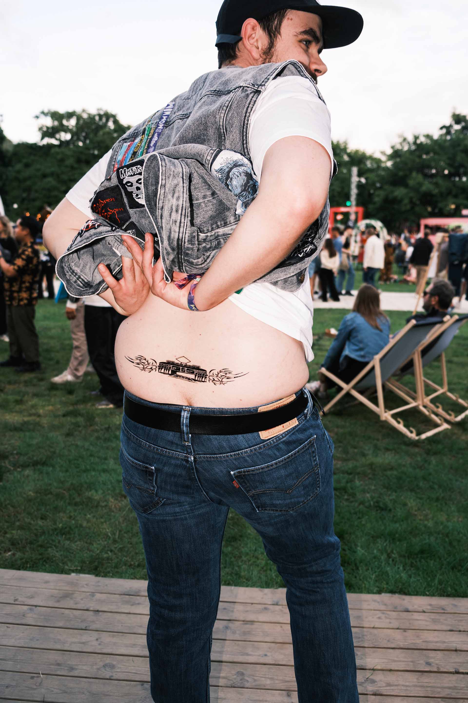 Person wearing a denim vest shows a tattoo of a tram with flame designs on their lower back at an outdoor event.