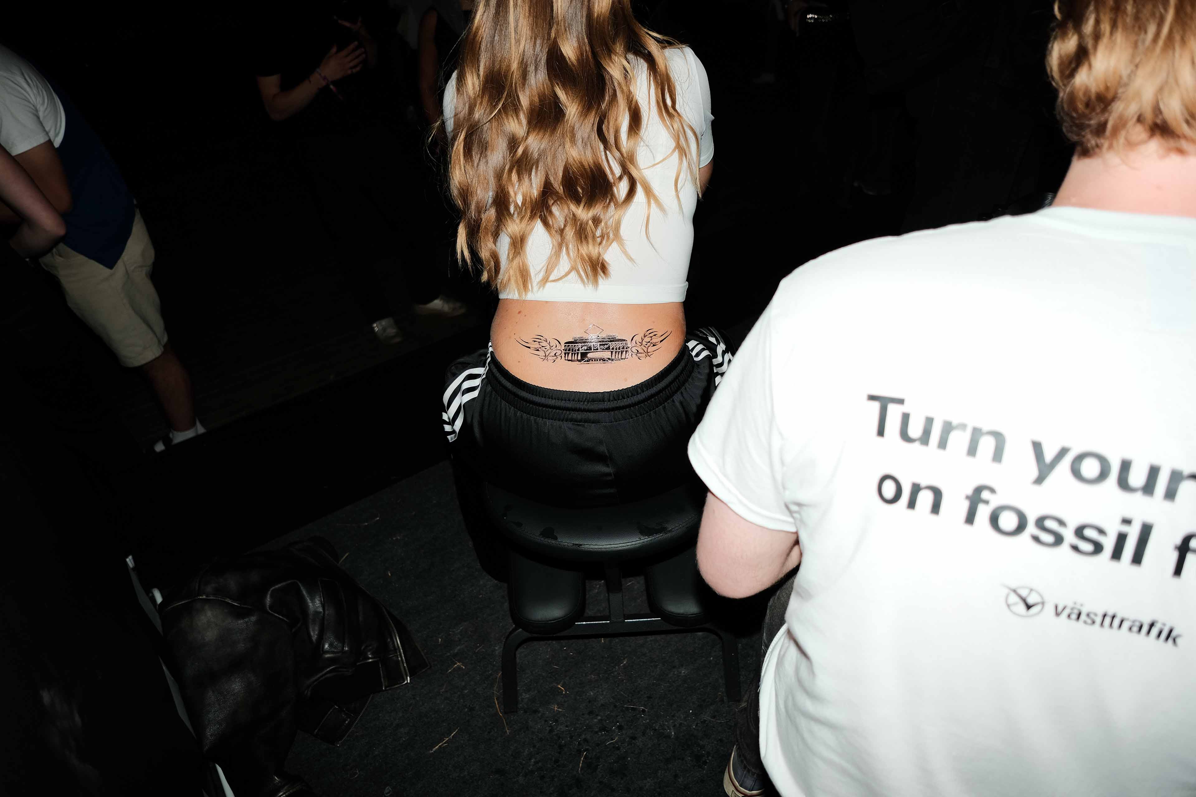 Woman with long wavy hair sitting on a stool, showing a tattoo of a tram with wings on her lower back, next to a person wearing a white shirt with partial text about fossil free.