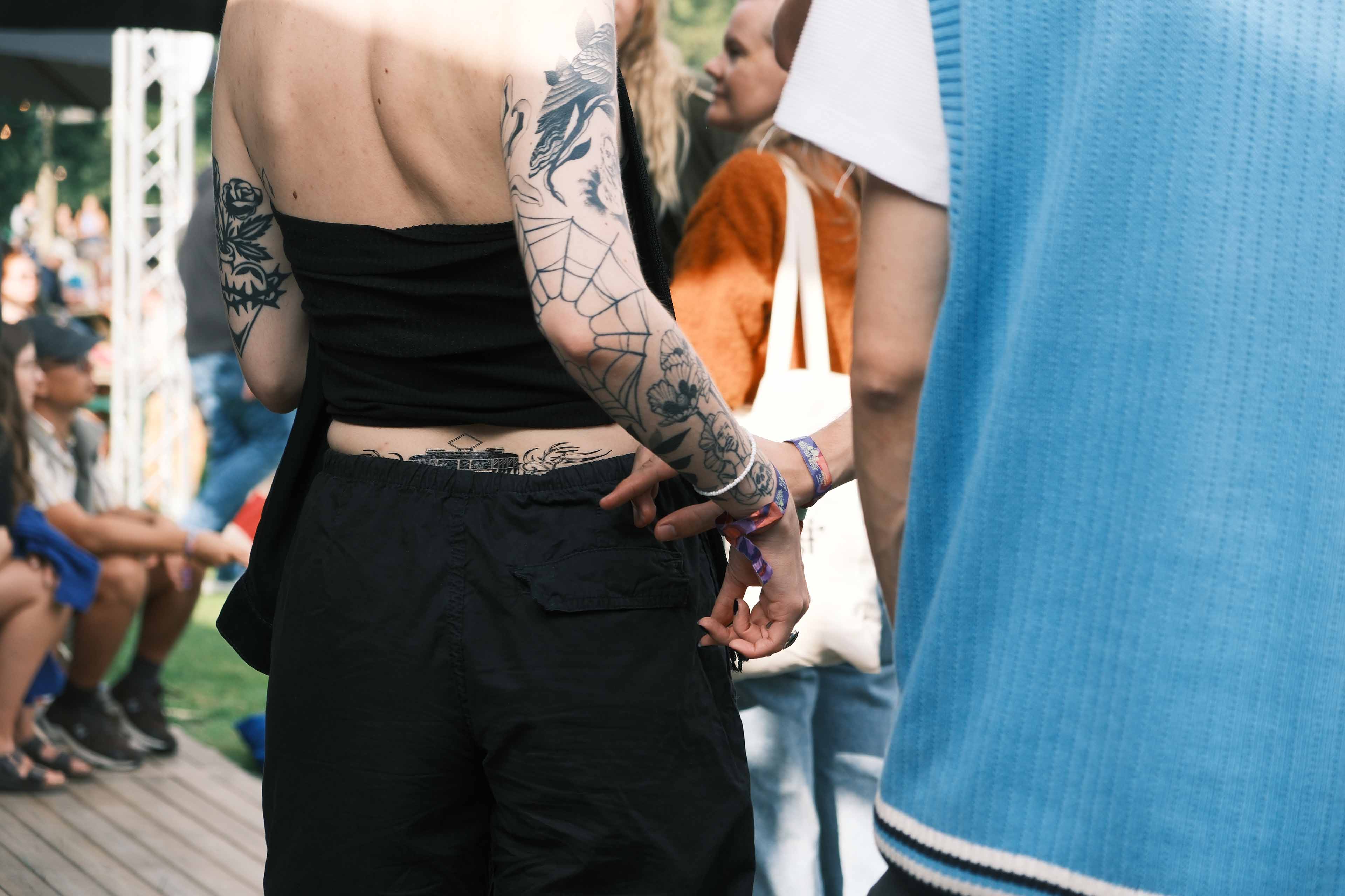 Person with multiple tattoos on arms and lower back wearing a black top and black pants, holding hands with another person.