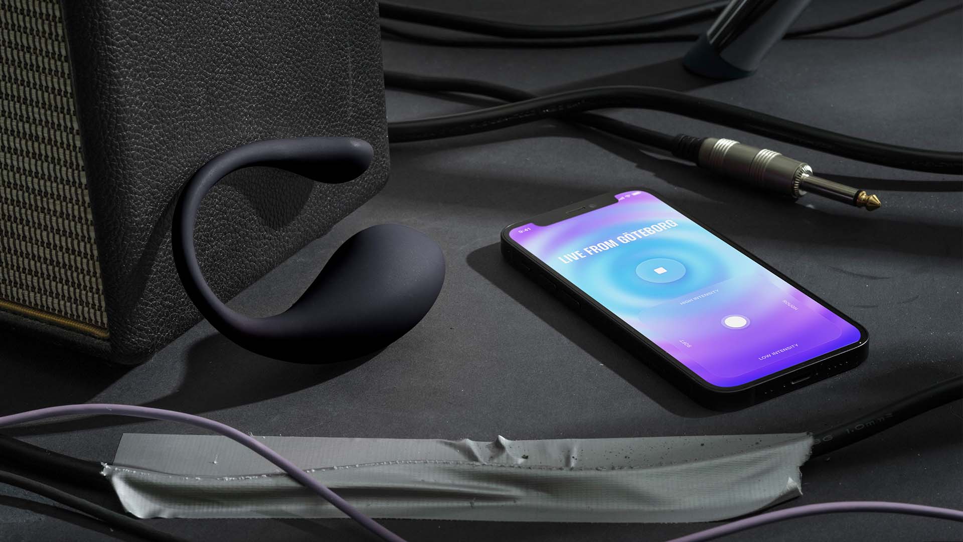 Black wearable device next to a smartphone displaying an app with 'Live from Göteborg' text and a control interface on a gray surface with cables.