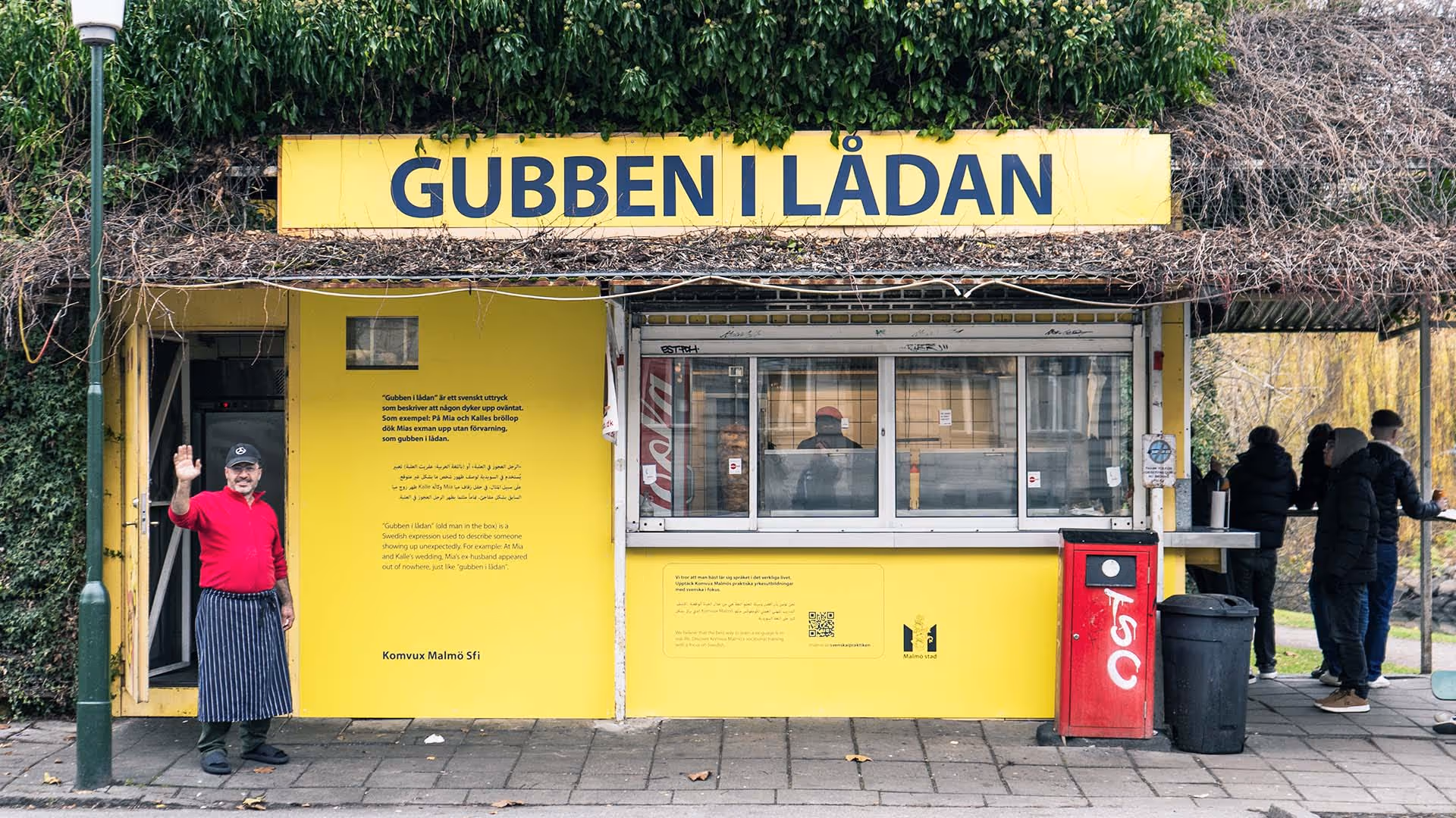 Man in red shirt and striped apron waving outside a yellow kiosk with sign 'GUBBEN I LÅDAN' and people standing at a nearby outdoor counter.