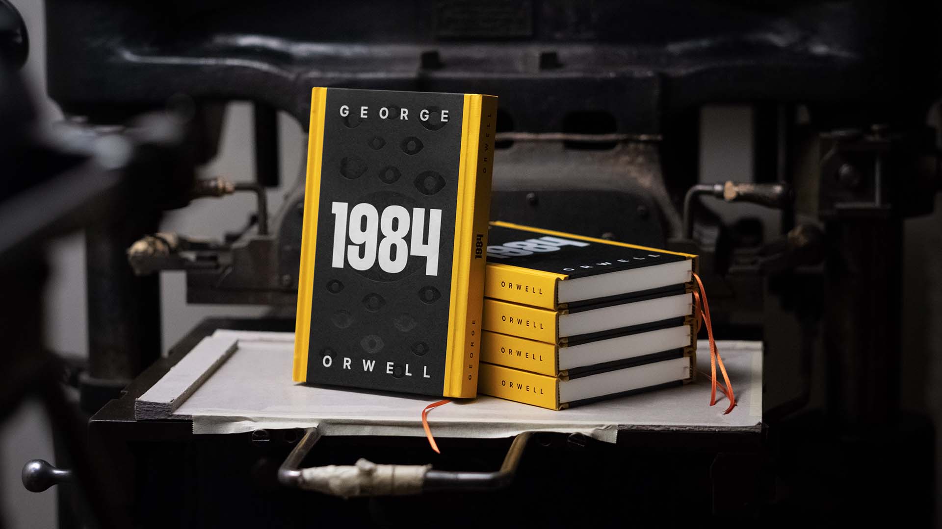 Stack of five hardcover books with yellow spines and black covers showing the title '1984' by George Orwell, placed on an old printing press.