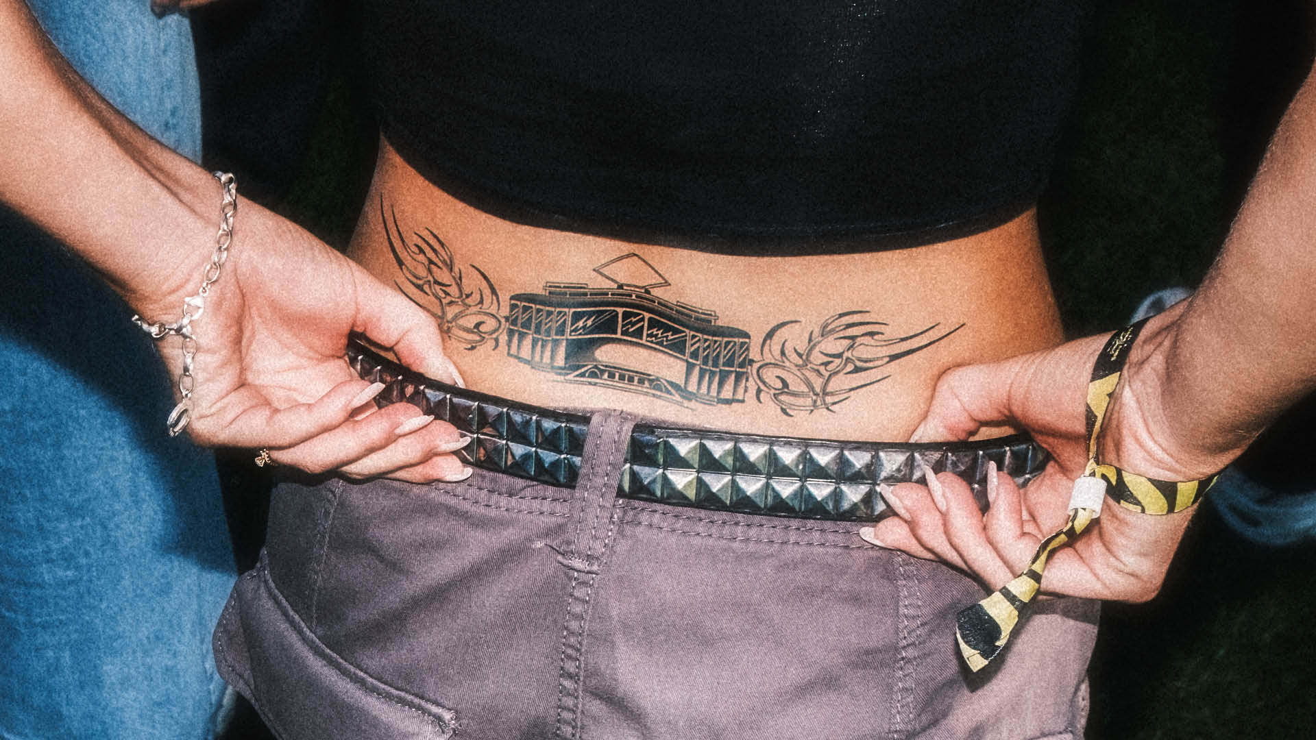 Close-up of a person's lower back showing a tattoo of a tram with tribal designs on either side, holding a studded belt.