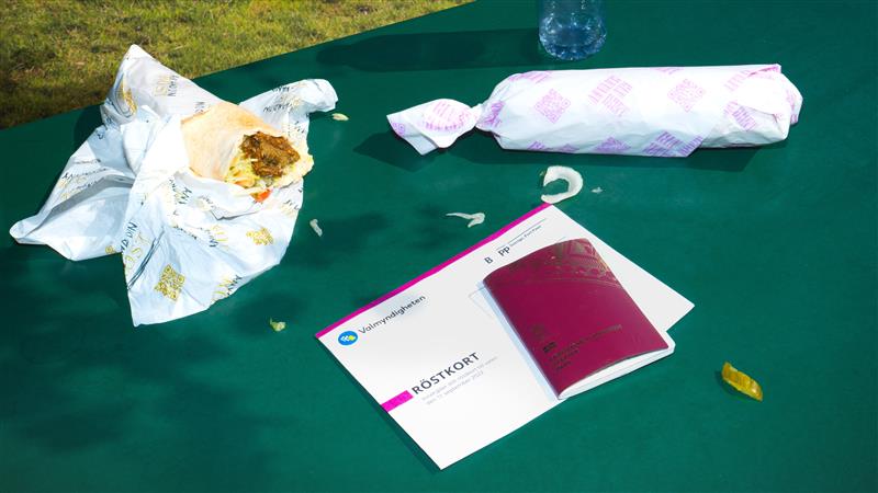 Partly unwrapped sandwich, wrapped food item, a water bottle, and a Swedish voting card with a maroon passport on a green table outdoors.