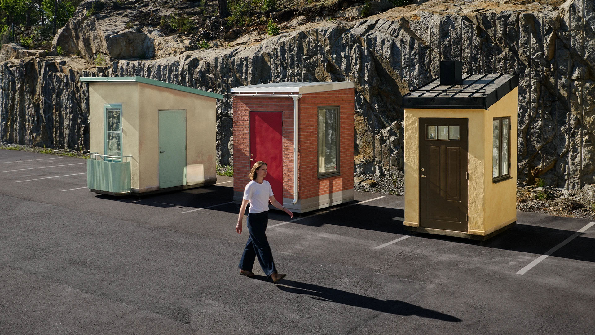A woman walking on asphalt past three small buildings in a row against a rocky hillside.